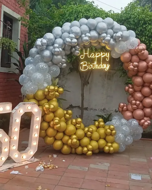 This outdoor setup for a 50th birthday party features a large circular balloon arch in silver, gold, and rose gold. The design is completed with a 'Happy Birthday' neon sign and large marquee numbers, perfect for garden or backyard celebrations.