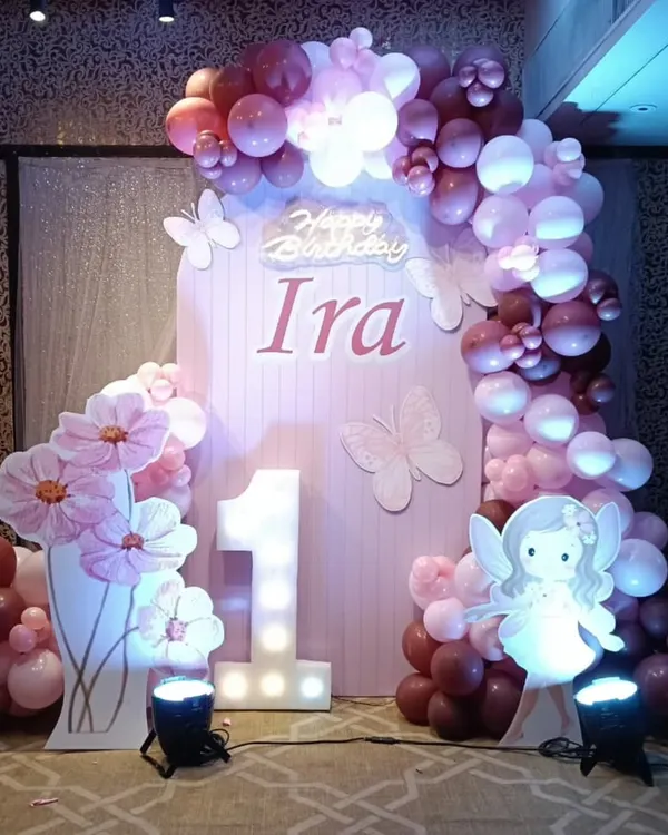 A beautiful fairy-themed first birthday for baby Ira. This setup includes a custom pink backdrop, a marquee number '1', a fairy cutout, and an organic balloon garland in shades of pink and purple.