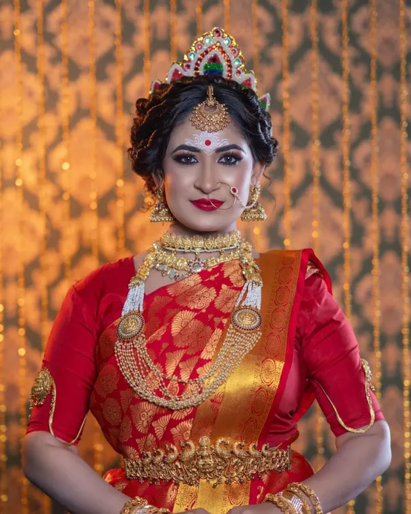 A classic Bengali bride, complete with the traditional 'Mukut' (crown), intricate white and red bindi design, and bold, kohl-rimmed eyes. The red Banarasi saree and gold jewellery complete this timeless look.