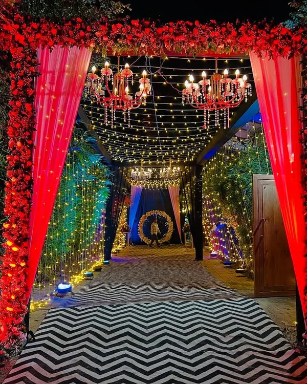 A spectacular entrance tunnel for an evening event. The combination of a dense red floral arch, hanging chandeliers, and a fairy light passage creates a grand and memorable welcome.