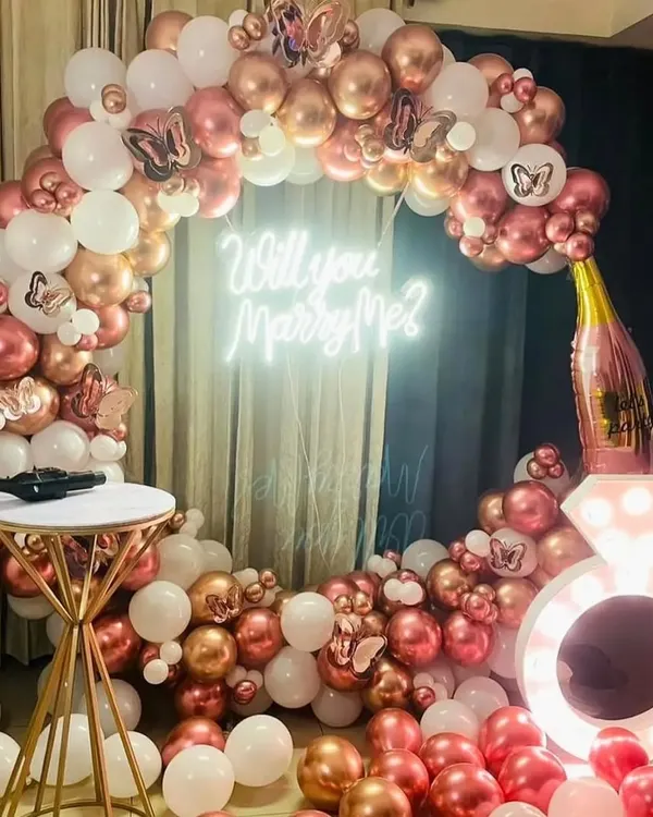 The perfect setup for a romantic proposal. This circular arch of rose gold and white balloons, complete with a 'Will you Marry Me?' neon sign and a light-up ring prop, creates a beautiful and memorable moment.
