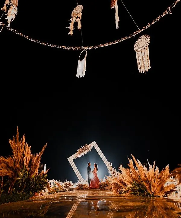 A breathtaking engagement setup under the stars. The couple is framed by a beautiful floral arch, surrounded by pampas grass and dreamcatchers, creating a truly magical "she said yes" moment.