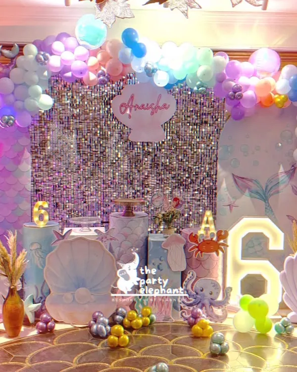 A stunning, wide view of our "Mermazing" celebration. This setup features multiple backdrops, a shimmering silver curtain, and an array of underwater props like shells, coral, and a light-up number '6'.