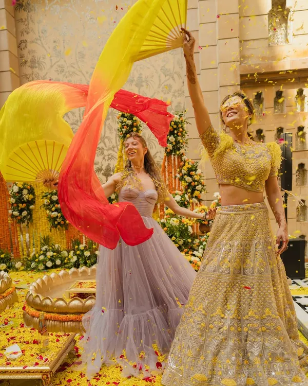 A Haldi ceremony is a festival of joy. Here, the bride and her friend dance with colorful fans amidst a shower of flower petals, capturing the high-energy and vibrant spirit of the event.