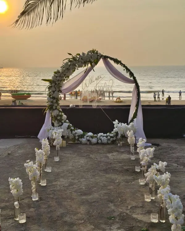 A breathtaking beach proposal I designed in Mumbai. The setup features a circular floral arch with white flowers and delicate draping, a candlelit aisle, and a custom 'Will you Marry Me?' sign, all timed perfectly for a golden sunset.