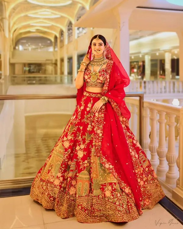 A full-length shot of the bride in a magnificent red lehenga, taken at the Omaxe Chowk. The grand, well-lit architecture of the location provides a stunning backdrop that enhances the regality of the bridal attire.