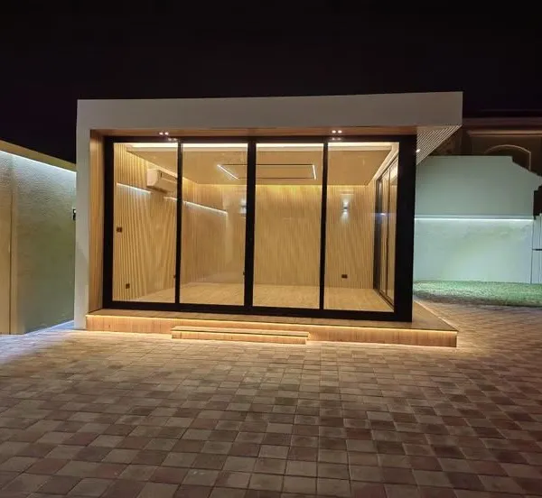A modern, standalone glass cabin illuminated at night, creating a striking visual. This custom backyard office pod features a black metal frame, warm wood-paneled interiors, and integrated LED lighting along the base, showcasing a premium and contemporary design.