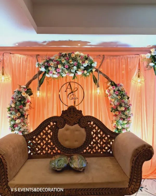 A classic engagement stage design featuring an ornate, carved wooden sofa for the couple. The backdrop is a soft peach drape with a beautiful floral arch and a custom monogram of the couple's initials, all lit by warm, hanging lights.