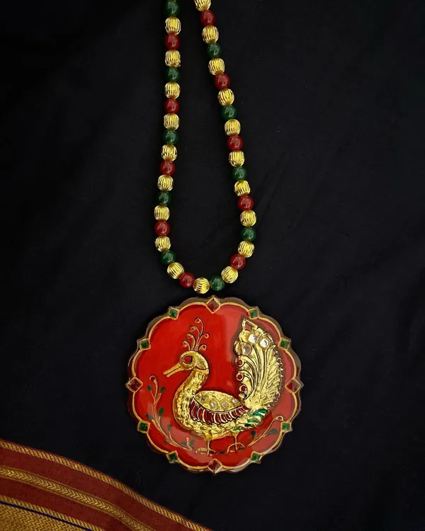 A stunning Tanjore art pendant featuring a peacock motif, crafted with authentic gold foil on a deep red base. The piece is finished with a protective resin coating, ensuring its lasting beauty and shine.