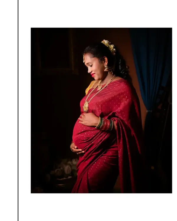 This portrait uses dramatic, low-key lighting to create a sense of intimacy and reverence, reminiscent of a classic painting. The focus is on the mother's gentle expression as she cradles her bump, adorned in a beautiful red silk saree and traditional jewelry.