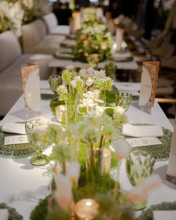 A long-table view of our green and white floral decor concept. The arrangement of fresh flowers, moss placemats, and elegant glassware creates a cohesive and breathtaking dining landscape for a large social gathering.