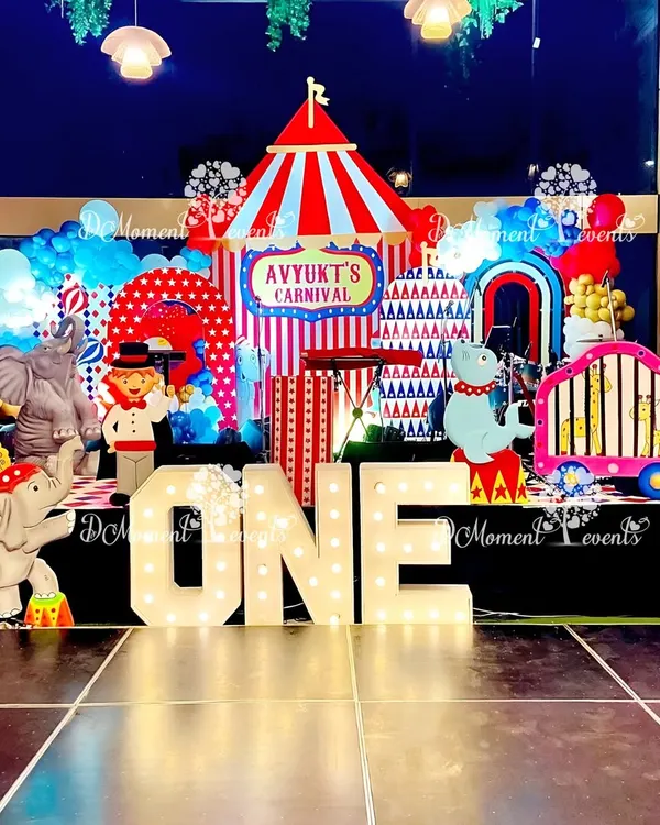 A spectacular carnival-themed stage for a first birthday, featuring a big top tent backdrop, animal props in a circus train, and large light-up marquee letters.