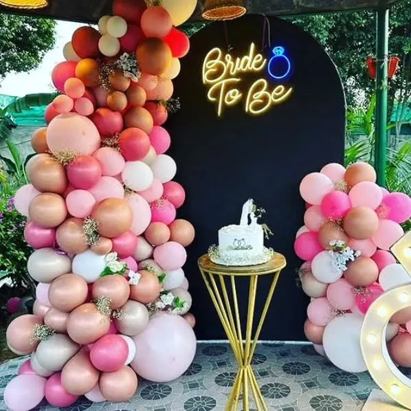 This chic bridal shower backdrop features a black arch panel with a 'Bride To Be' neon sign. It's framed by a lush organic balloon garland in shades of pink, nude, and white, with a small pedestal for the cake.