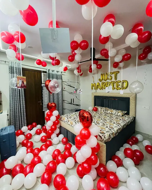 A classic "Just Married" bedroom decoration. The room is filled with red and white balloons on the floor and ceiling, with foil letters above the bed and personalized photos hanging from ribbons for a sweet, memorable touch.