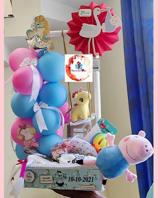 A cheerful baby arrival hamper with a gender-neutral color scheme. It features a Peppa Pig plush, balloons, baby clothes, and toys, all creatively arranged.