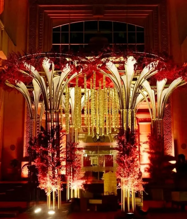 A breathtaking entrance for a grand reception, featuring a massive, sculptural structure adorned with red flowers and hanging golden strings, all lit to perfection.