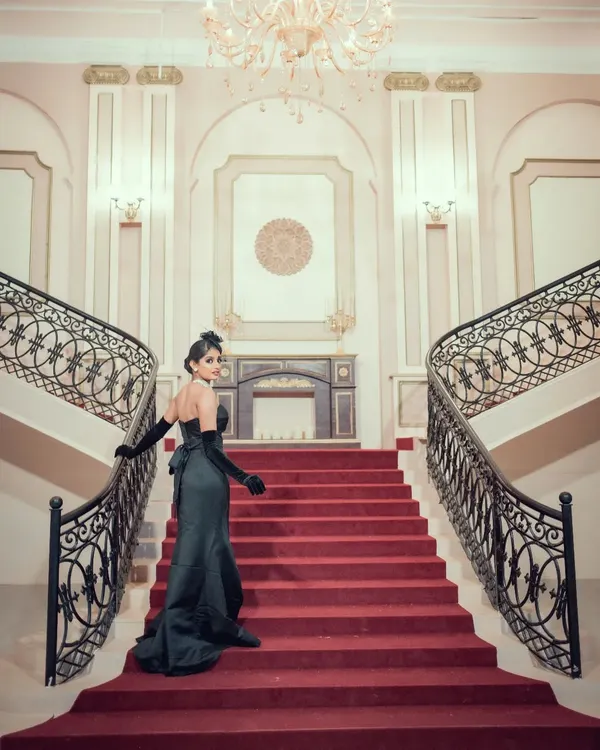 A grand, cinematic shot of a woman in an elegant black gown on a red-carpeted staircase. This image evokes the glamour and drama of old Hollywood, perfect for a vintage-themed shoot.