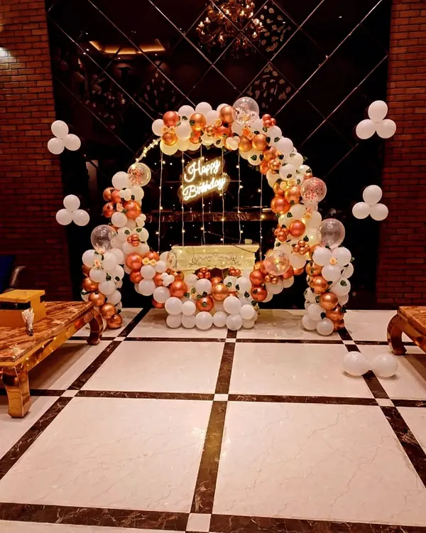 A beautiful birthday ring decoration with white and rose gold balloons, a neon sign, and fairy lights, creating an elegant photo spot.