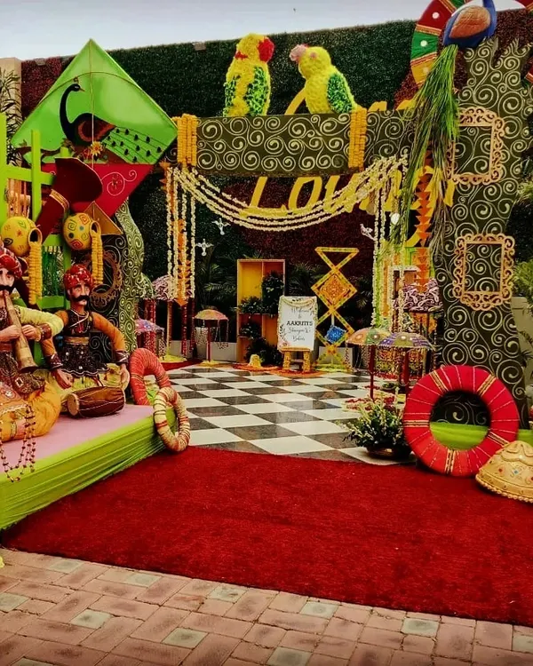 A full-scale Teej festival setup, rich with Rajasthani cultural elements. This photo corner features kathputli puppets, colorful kites, floral peacocks, and a checkered floor, creating a vibrant and traditional celebratory vibe.