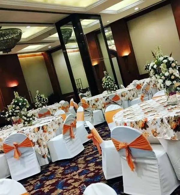 This is a full view of a banquet hall I decorated for a day function. The setup includes round tables with printed cloths, orange bows on the chairs, and fresh floral centerpieces on each table.