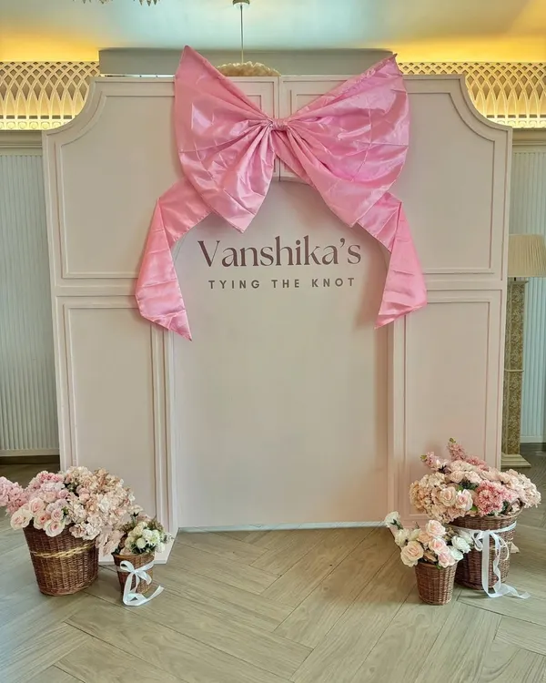 A chic backdrop for "Vanshika's Tying the Knot" bridal shower. The design features a large pink bow on a paneled wall, with baskets of pink roses completing the feminine and celebratory theme.