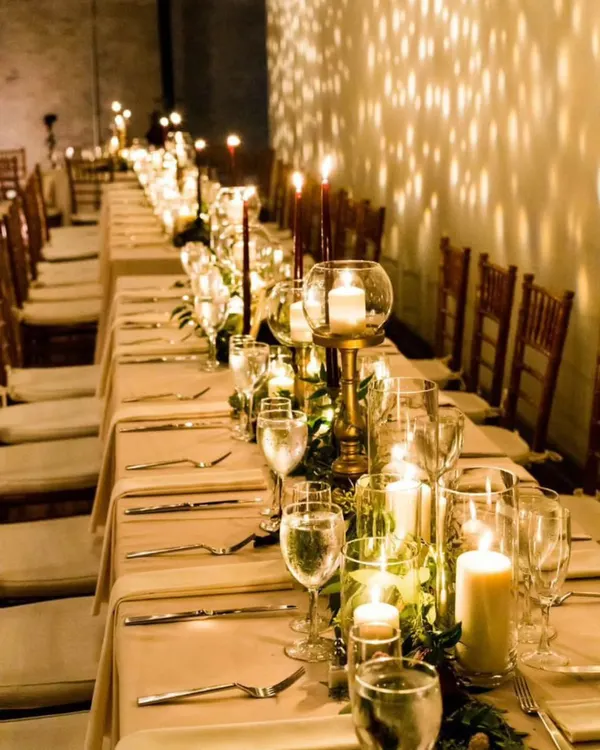 This long dining table is set for a formal evening event, illuminated entirely by candlelight. The warm glow from numerous pillar and taper candles creates a dramatic and intimate atmosphere, reflecting off the glassware.
