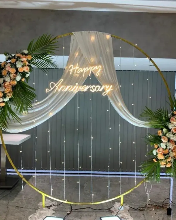 This elegant anniversary backdrop features a golden circular frame, a custom neon sign, delicate fairy lights, and fresh floral arrangements. It’s a sophisticated choice that creates a perfect focal point for celebrations in banquet halls or at home.