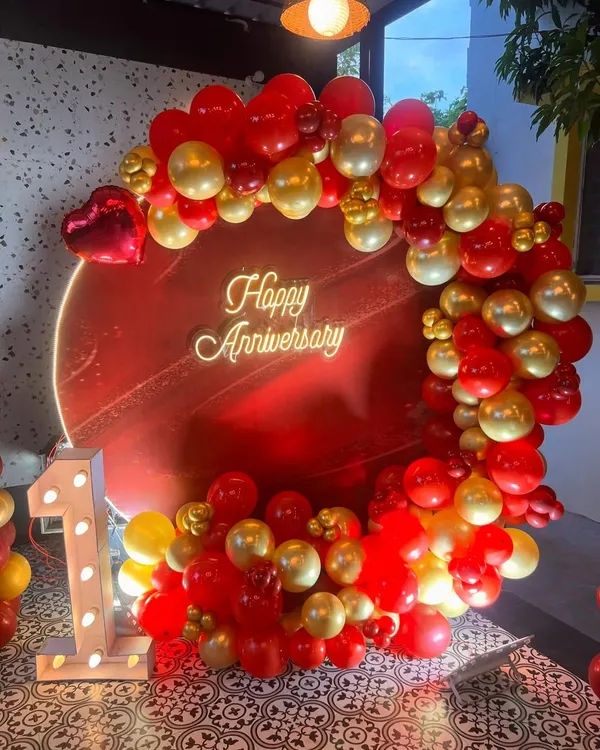 I design bold and beautiful backdrops to celebrate your special milestones. This first-anniversary setup features a vibrant red circular panel, a custom 'Happy Anniversary' neon sign, and a lush garland of red and gold balloons, complete with an illuminated number '1'.
