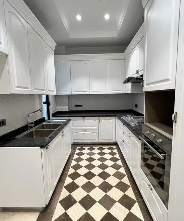 A full view of a Victorian classical kitchen. It features white PU-finished cabinets, a black granite countertop, and a striking checkerboard floor. We include a built-in oven and hob, ensuring the space is ready to use.