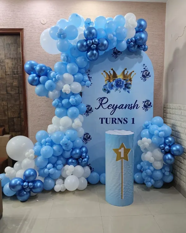 A royal prince theme for a first birthday. This beautiful setup features a custom blue backdrop with a crown motif, an organic balloon arch in shades of blue and white, and a themed pedestal.