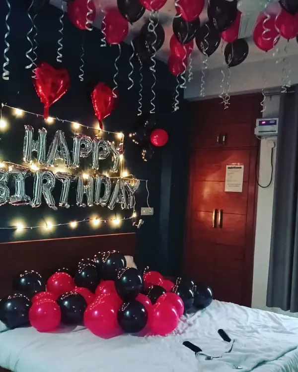 A classic birthday bedroom surprise. The setup includes black and red balloons on the bed and ceiling, with 'Happy Birthday' foil letters illuminated by warm fairy lights on the wall.