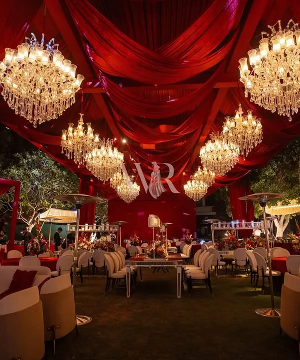 This is the essence of a royal darbar fantasy. The entire venue is enveloped in deep crimson drapes, with a ceiling full of chandeliers casting a golden glow on the plush, elegant seating arrangements below.