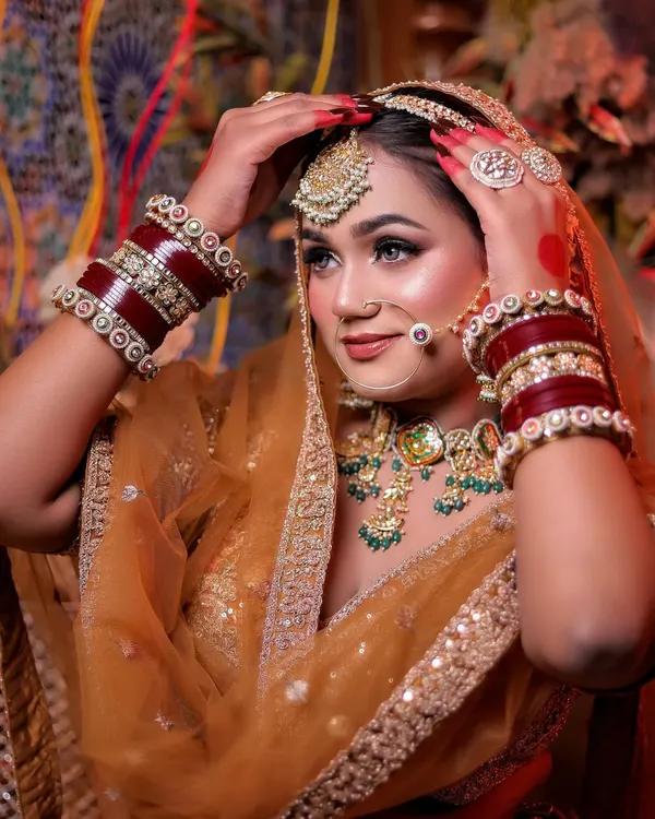 A beautiful close-up of a bride in a mustard-gold lehenga, adjusting her dupatta. The red chooda bangles add a pop of traditional color.