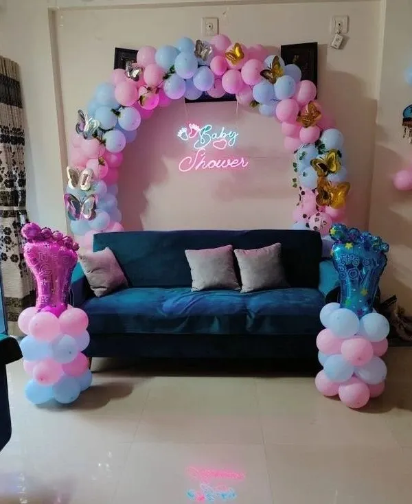 This is a great example of a compact baby shower setup perfect for a living room. It features a balloon arch over the sofa, a "Baby Shower" neon sign, and fun foil balloons shaped like baby feet to complete the look.