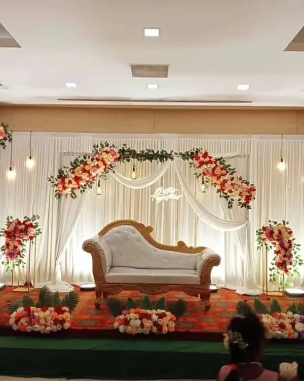 I designed this elegant stage for an intimate reception in a small banquet hall. The setup features a classic chaise lounge, soft white drapery, and beautiful artificial floral arrangements, all brought together with warm, hanging Edison bulbs for a romantic ambiance.