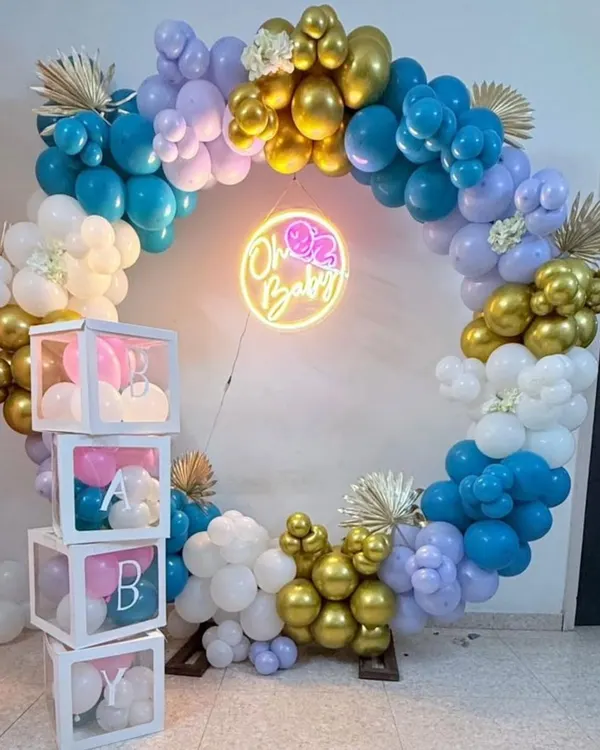 A modern baby shower backdrop featuring a full balloon ring in shades of blue, purple, and gold. The "Oh Baby" neon sign adds a warm glow, and the stacked 'BABY' blocks complete this popular party setup.