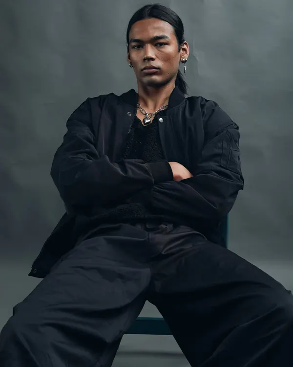 A powerful, seated portrait with a moody, grey background. The model's crossed arms and direct gaze convey a sense of strength and confidence. The lighting is soft yet dramatic, sculpting his features.