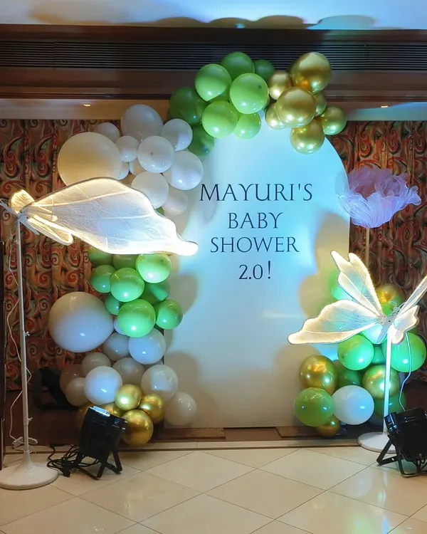 A modern and minimalist baby shower setup with a green and gold balloon garland on a white arched backdrop. The look is completed with illuminated butterfly props and wooden 'BABY' blocks.