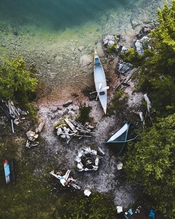 An aerial view of a complete lakeside setup. You can see the tent, the central campfire ring, and a canoe ready for exploration. I choose locations that offer both relaxation and the opportunity for gentle adventure on the water.