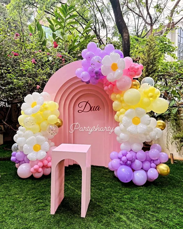 A beautiful outdoor cake cutting setup with a pink arched backdrop and daisy-themed balloon decorations on a lush green lawn.