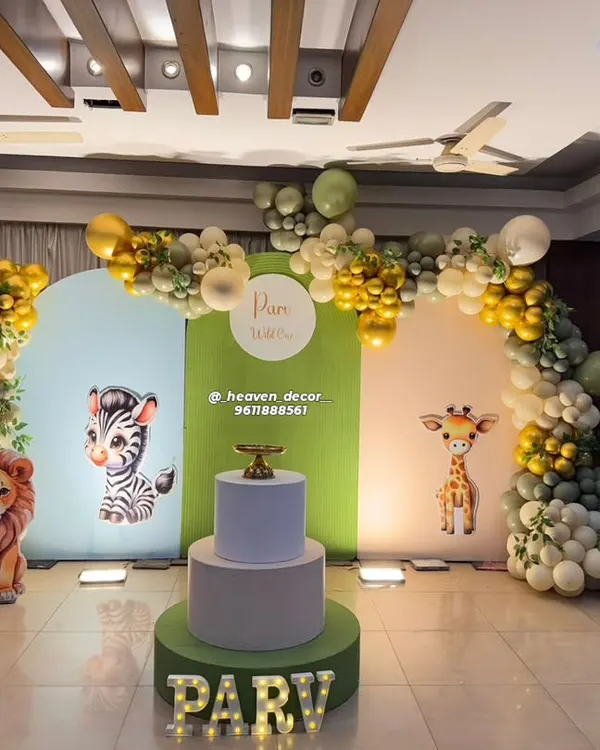 A classic "Wild One" jungle theme for a first birthday, using a three-panel arched backdrop with cute safari animal graphics. The scene is framed by an organic balloon arch in gold and shades of green, with marquee letters for the name.