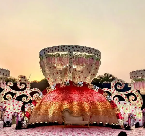 A truly unique reception stage design featuring massive, ornate structures covered in a gradient of red and pink flowers. The central floral dome creates a dramatic focal point for the couple's seating, perfect for a lavish outdoor celebration.