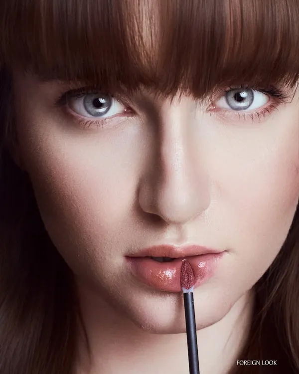 A classic cosmetics beauty shot. The focus is on the glossy lip product being applied, with the lighting perfectly capturing the texture and color while keeping the model's eyes bright and engaging.