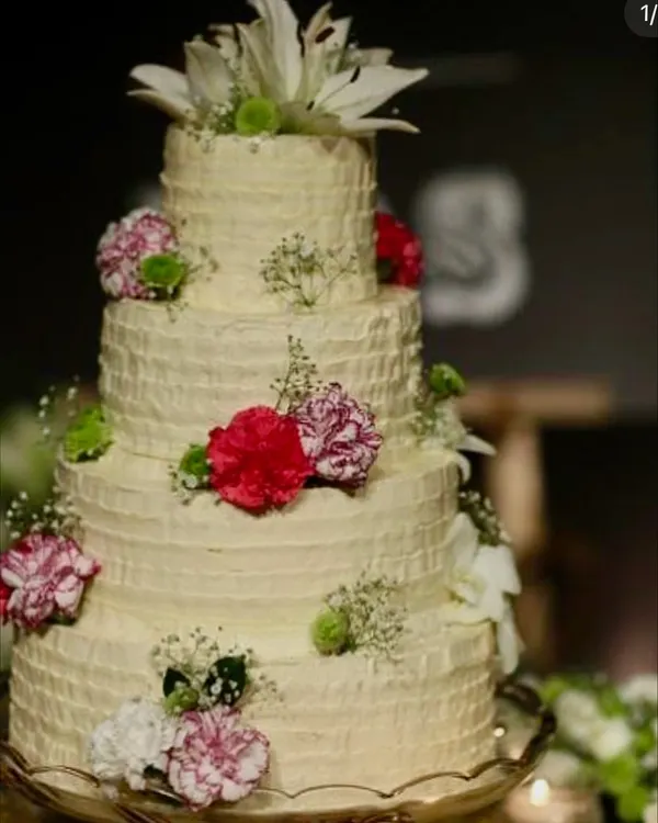 A magnificent four-tiered wedding cake with a rustic, textured buttercream finish, decorated with fresh carnations, lilies, and baby's breath. This large celebration cake is designed to be a showstopper at any grand wedding reception.