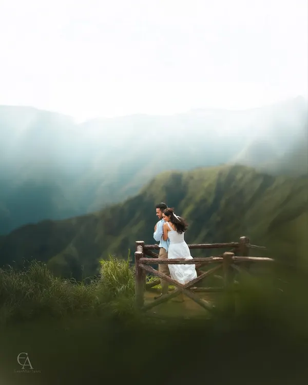 A couple shares an intimate moment on a wooden viewpoint, overlooking a breathtaking valley of green mountains. The soft mist adds a dreamy quality to this stunning landscape portrait.