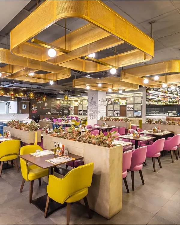 A wide view of the Pasta Street restaurant, showcasing the custom-designed network of golden perforated sheet lighting that creates a warm and inviting canopy over the central dining area. The vibrant yellow and pink seating adds a playful yet chic energy to the space.
