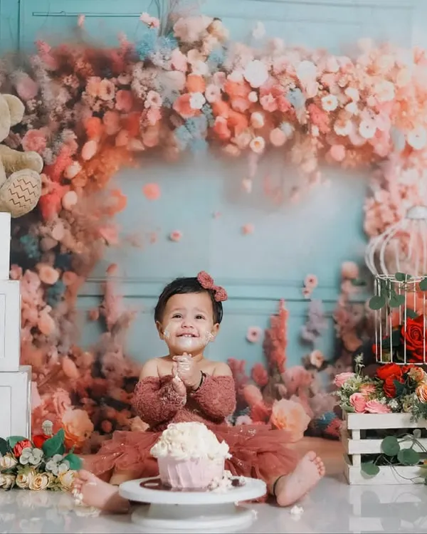 A perfect first birthday celebration. This little girl is all smiles, ready to dig into her cake in a stunning studio setup featuring a floral arch, teddy bears, and 'ONE' blocks.