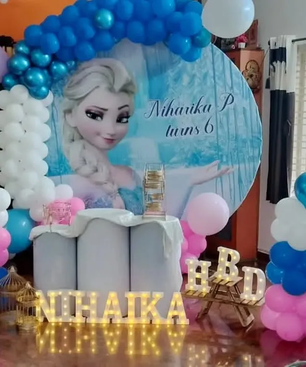 A magical Frozen-themed birthday party for Niharika's 6th birthday. The centerpiece is a large circular backdrop featuring Elsa, framed by a blue, white, and pink balloon arch, perfect for a home celebration.