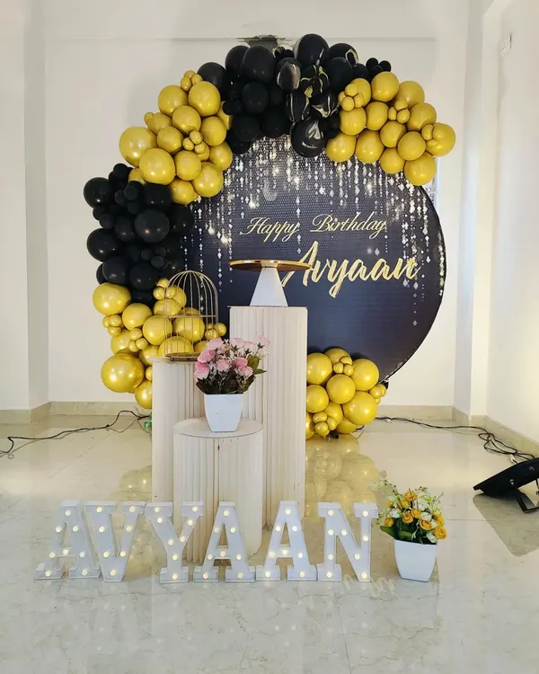 A chic and modern birthday setup with a black and gold balloon arch. This decoration includes a personalized backdrop with a starlight effect, elegant white plinths, and marquee letters for the name.