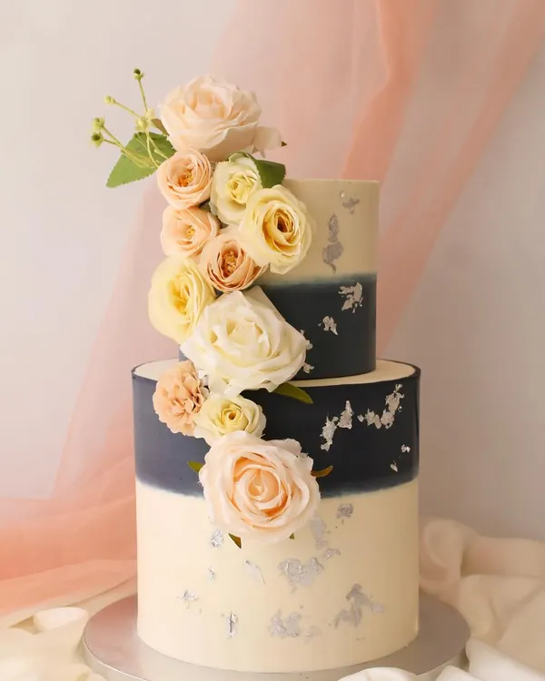 A modern two-tier cake featuring a navy blue to white ombre effect, accented with delicate silver leaf and a cascade of beautiful peach and cream roses. This elegant design is perfect for weddings, engagements, or sophisticated celebrations.